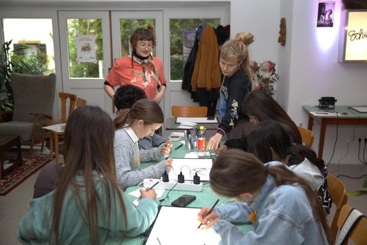 Schüler/innen zeichnen an Tisch auf Papier und Tablets, zwei Frauen im Hintergrund