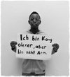 Schwarzweiß-Foto eines Schülers, der ein Schild mit der Aufschrift „Ich bin Kongoleser, aber bin nicht arm“ hochhält.