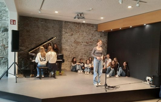 eine Schülerin singt auf Bühne, zwei spielen auf Flügel, weitere stehen und sitzen im Hintergrund