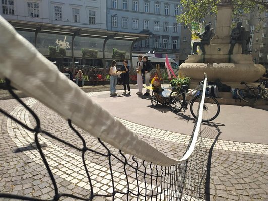Foto von einem öffentlcihen Platz vor einem Brunnen. Ein Tennisnetz ist gespannt. 