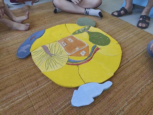 Kinder sitzen vor einem Holzpuzzle mit einem gezeichneten Haus und zwei Bäumen