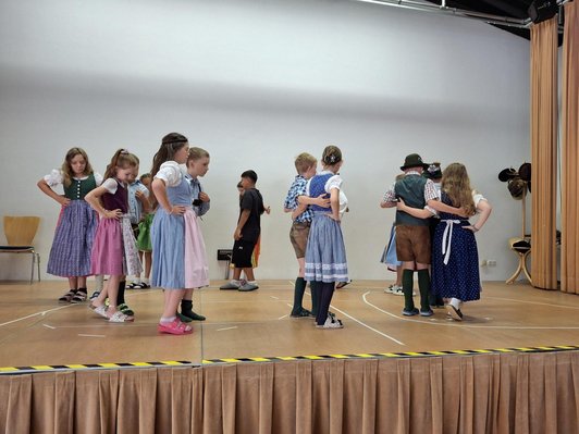 Kinder in Lederhosen und Dirndl tanzen auf einer Bühne.