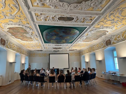 Schüler/innen sitzen und Mann steht in Saal mit prunkvoller Stuckdecke im Kreis