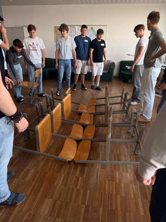 Schüler stehen um Stühle die am Boden liegen und zueinander schauen