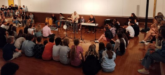 Schüler/innen sitzen um eine Musikgruppe (Ensemble) aus Schüler/innen und Musiker/innen