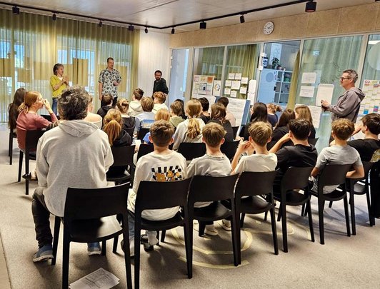 Schüler/innen sitzen Reihen vor und neben vier Erwachsenen