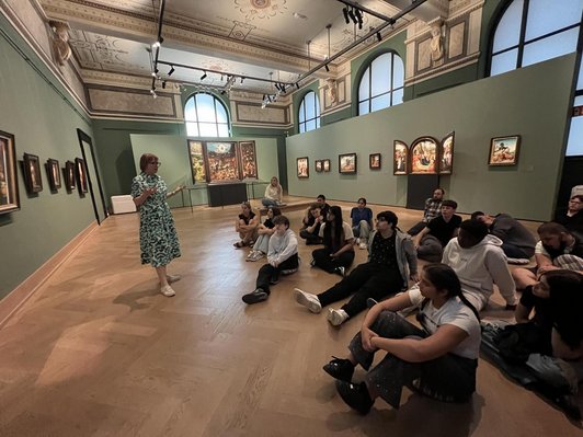 Schüler/innen sitzen in Museumsraum am Boden und blicken auf stehende Frau