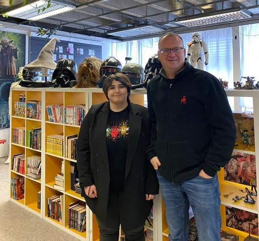 Eine Referentin und ein Referent stehen vor einer Bücherstellage in einer Bibliothek