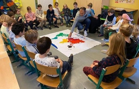 Volksschulkinder sitzen im Sesselkreis und Workshopsleiter erklärt eine Grafik einer bunten Weltkarte