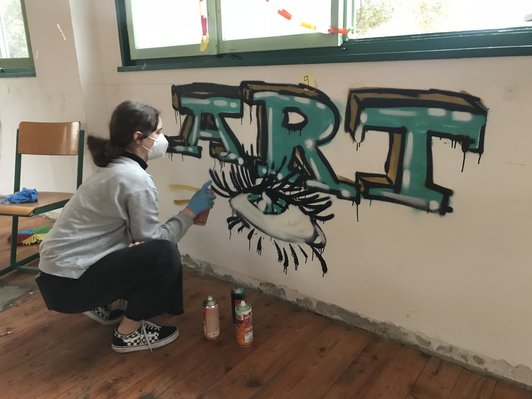 Foto einer Schülerin mit Schutzmaske, die hockend ein Graffiti mit dem Schriftzug ART und einem Auge auf eine Innenwand sprayt