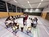 In einem Turnsaal ist ein Sesselkreis mit Schüler/innen, die alle vor sich eine Trommel haben. In der mItte stehen zwei Mädchen und halten die Trommel in die Höhe.