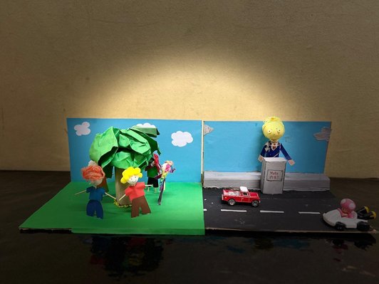 Foto von zwei Szenen. Erste von gebastelten Figuren im Kreis stehend um einen Papierbaum herum. Zweite Szene Figur hinter Rednerpult mit Schriftzug vote me! Dovor eine Straße mit zwei Autos.