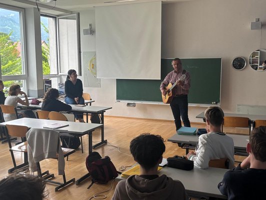 Schüler/innen und Lehrerin sitzen in Klassenzimmer und blicken auf Gitarre spielenden Mann