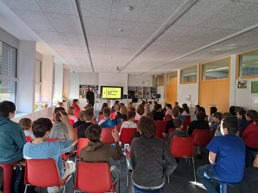 Schüler/innen sitzen in Reihen vor Mann und Präsentationsmonitor