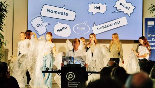 Foto von mehreren Schüler:innen auf der Bühne mit transparenten Foliern, ein Schüler sitzt im Vordergrund an einem Tisch mit Mikrophon und einem Unseco Papier in der Hand. Auf der Leinwand im Hintergrund steht auf verschiedene Sprachen Hallo.