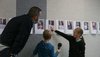 Foto einer Pinnwand mit Porträts von Schüler/innen; davor stehen eine Schülerin, die auf ihr Porträt an der Wand zeigt, und ein Schüler, der ein Aufnahmegerät in der Hand hält, sowie ein Erwachsener