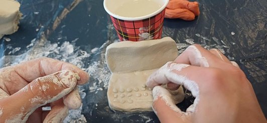 Zwei Hände mit Ton verschmiert basteln an einem kleinen Laptop aus Ton, dahinter ein Becher mit Wasser