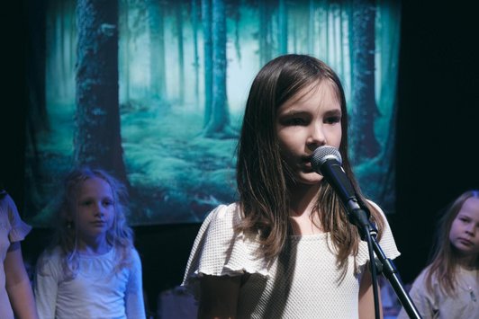 junge Schüler/in singt in ein Mikrophon, dahinter weitere Schüler/innen in einer Waldprojektion