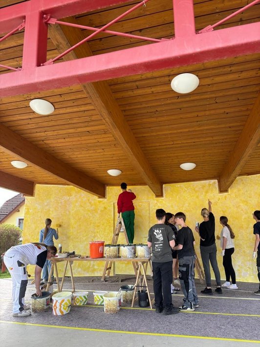 Foto vor einer gelben Wand mit Pinseln in der Hand. Im Vordergrung gelbe Farbkübel. Schüler:innen streichen die Wand.