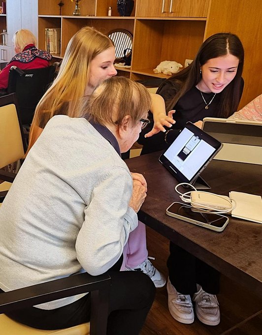 zwei Mädchen und eine Seniorin blicken an Tisch sitzend auf Teblats