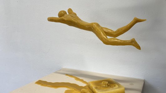 Gelbe Figur aus Wachs geformt die über einem geschmolznem Wachsstück schwebt.
