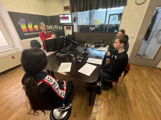 Drei Schüler/innen sitzen in einem Radiostudio an einem Tisch und lesen Texte in ein Mikro. Die Sendung wird von einer Vermittlerin betreut, die vor Ihnen steht.