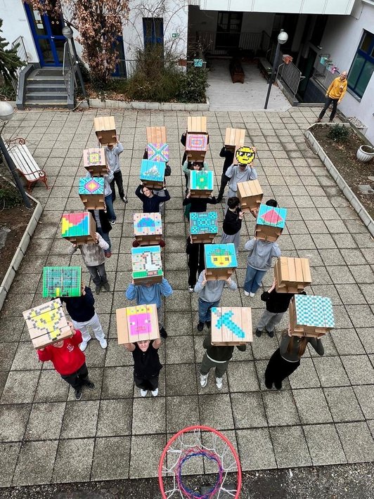 Foto von Schüler:innen mit quadratischen Kartonschachteln als Hocker mit unterschiedlichen Pixelbildern bemalt aus der Vogelperspektive.
