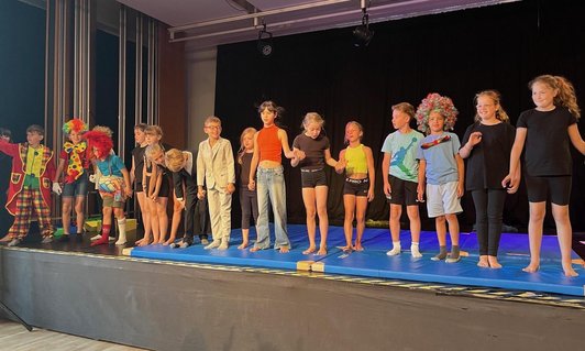 18 Kinder, teils in Clown-Kostümen, stehen Hände haltend in einer Linie auf einer Bühle