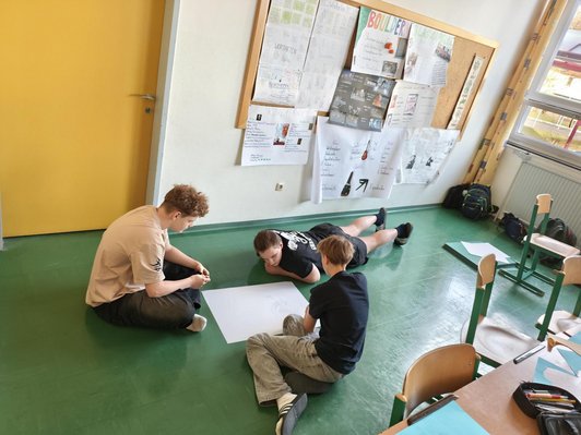 Foto von drei Schhülerm sitzend bzw. liegend am Boden im Klassenzimmer. In der Mitte aiengroßes Blatt Papier.