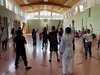 Kinder stehen in einem Turnsaal mit dem Rückem zum Betrachter/ zur Betrachterin und heben die rechte Hand hoch. Sie zeigen mit einem Finger nach oben.