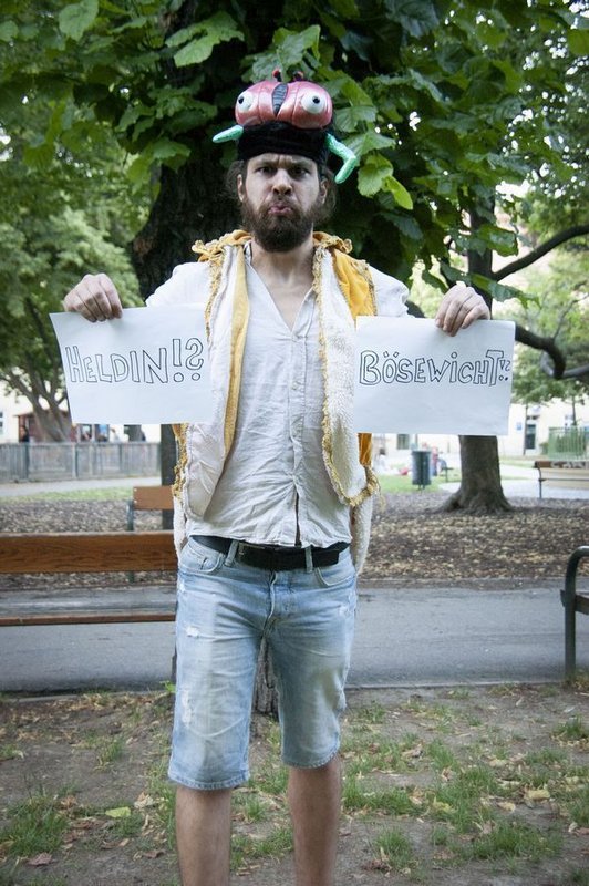 'Foto des Künstlers mit lustiger Kopfbedeckung in einem Park mit zwei Schildern in den Händen, auf denen ''Heldin!?'' und ''Bösewicht!?'' steht '